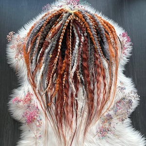 Puede incluir: Una colección de rastas hechas a mano en tonos naranja, marrón, gris y blanco. Las rastas están dispuestas sobre una alfombra de piel sintética blanca y decoradas con pequeñas flores de colores. Las rastas son una mezcla de texturas y estilos.