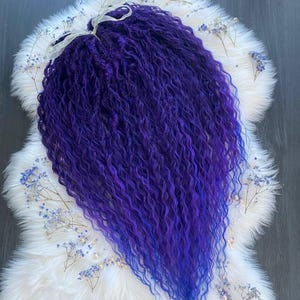 May include: A bundle of long, wavy, purple synthetic hair extensions. The hair transitions from a deep purple at the roots to a vibrant blue at the ends. The extensions are arranged on a white, fluffy surface, decorated with small, blue flowers.