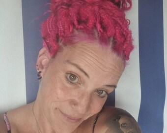 Op de afbeelding: Een persoon met felroze krullend haar in een hoge knot, met een dunne zilveren ketting met een kleine sterhanger, een neuspiercing en zwarte oorpiercings. Een realistische tatoeage van een hondenportret is zichtbaar op de bovenarm.