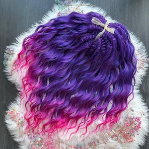 May include: A bundle of wavy, ombre hair extensions in shades of purple and pink. The hair transitions from a deep purple at the roots to a vibrant pink at the ends. A light beige ribbon secures the top of the bundle. The hair is placed on a white, fluffy surface.