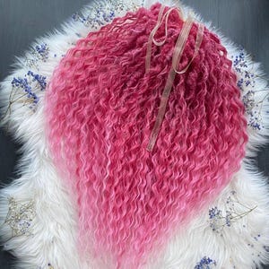 Puede incluir: Una peluca rizada rosa con un degradado de color, que va desde un magenta intenso en las raíces hasta un rosa suave en los extremos. La peluca se muestra sobre una superficie blanca y esponjosa, con pequeñas decoraciones florales azules.