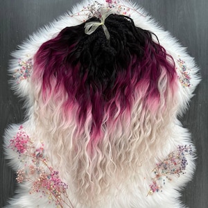 May include: Black, burgundy, pink, and white ombre hair extensions with a wavy texture. The extensions are arranged on a white faux fur rug, decorated with small, colorful flowers and a yellow ribbon.