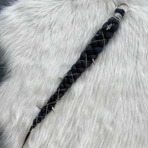 Puede incluir: Extensión de cabello trenzado negro con detalles plateados, incluyendo cadenas y púas metálicas. La extensión presenta un anillo plateado en la parte superior y un dije de ankh. Se muestra sobre una superficie blanca y esponjosa.