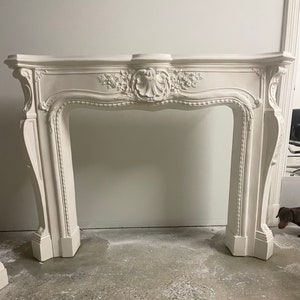 May include: White ornate fireplace mantel with intricate carvings and a scalloped design. The mantel is made of a smooth, white material and has a classic, elegant style.