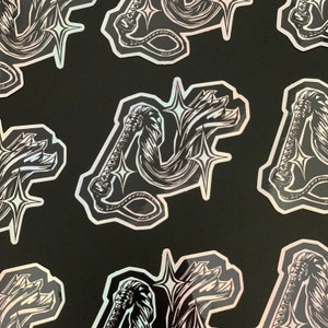 May include: A set of black and white holographic stickers featuring a stylized illustration of a whip with stars. The whip is coiled and has a handle with a knot at the end. The stars are stylized and have a pointed shape.