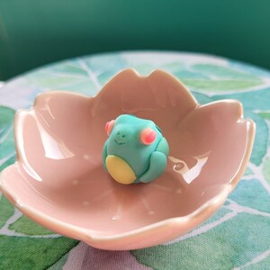 May include: A small, teal green frog figurine with pink eyes and a yellow belly, sitting in a pink ceramic dish.