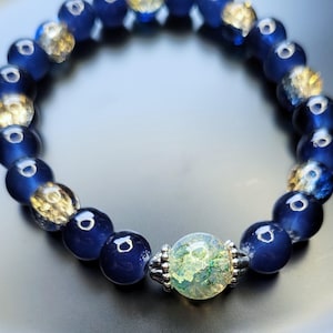 Könnte beinhalten: Ein blaues und klares Glasperlenarmband mit einer grünen und weißen Glasperle als Mittelpunkt. Das Armband besteht aus runden Perlen und hat einen silbernen Verschluss.