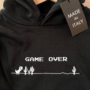 Könnte beinhalten: Schwarzer Kapuzenpullover mit weißem "GAME OVER"-Motiv mit einem pixeligen Dinosaurier und Kakteen. Der Kapuzenpullover hat eine Kapuze und ein Etikett mit der Aufschrift "MADE IN ITALY".