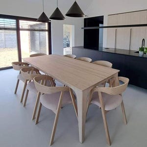 Puede incluir: Un comedor moderno con una mesa rectangular de madera clara y ocho sillas a juego. Las sillas tienen respaldos curvos y tapicería de color claro. Tres lámparas colgantes cónicas negras cuelgan sobre la mesa.