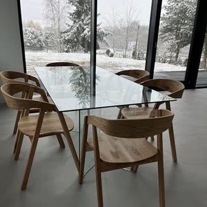 K&ouml;nnte beinhalten: Ein modernes Esszimmer-Set mit einem rechteckigen Glastisch und sechs Holzst&uuml;hlen. Die St&uuml;hle haben geschwungene R&uuml;ckenlehnen und abgewinkelte Beine. Der Raum hat gro&szlig;e Fenster mit Blick auf eine verschneite Au&szlig;enansicht.