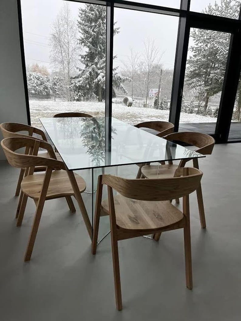 K&ouml;nnte beinhalten: Ein modernes Esszimmer-Set mit einem rechteckigen Glastisch und sechs Holzst&uuml;hlen. Die St&uuml;hle haben geschwungene R&uuml;ckenlehnen und abgewinkelte Beine. Der Raum hat gro&szlig;e Fenster mit Blick auf eine verschneite Au&szlig;enansicht.