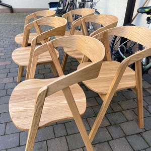 K&ouml;nnte beinhalten: Sechs Holzst&uuml;hle mit geschwungenen R&uuml;ckenlehnen und abgerundeten Sitzen. Die St&uuml;hle sind aus hellem Holz gefertigt und haben ein minimalistisches Design. Die St&uuml;hle stehen im Freien auf einem Backsteinpflaster.
