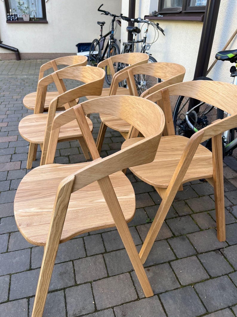 K&ouml;nnte beinhalten: Sechs Holzst&uuml;hle mit geschwungenen R&uuml;ckenlehnen und abgerundeten Sitzen. Die St&uuml;hle sind aus hellem Holz gefertigt und haben ein minimalistisches Design. Die St&uuml;hle stehen im Freien auf einem Backsteinpflaster.