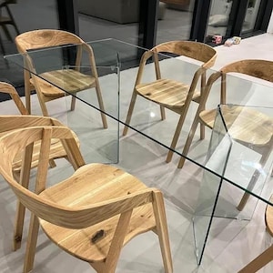 K&ouml;nnte beinhalten: Ein modernes Esszimmer-Set mit einem rechteckigen Glastisch und sechs Holzst&uuml;hlen. Die St&uuml;hle haben geschwungene R&uuml;ckenlehnen und Beine, und der Tisch hat eine transparente Glasplatte. Das Holz hat eine nat&uuml;rliche Oberfl&auml;che.