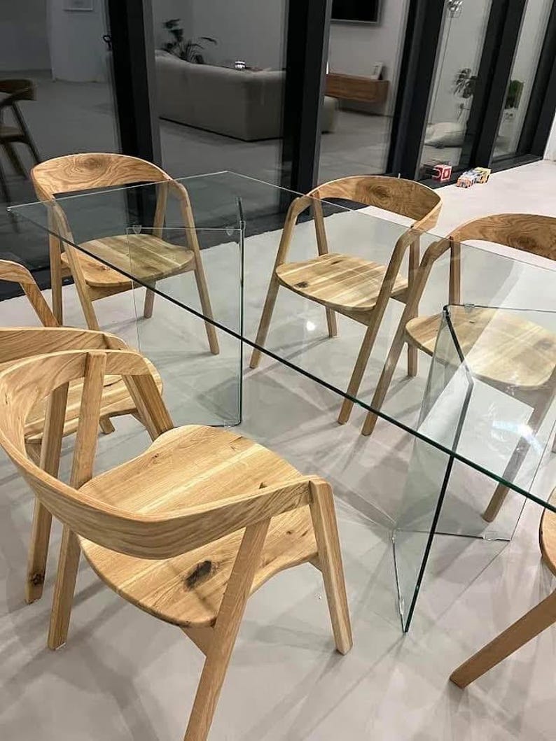 K&ouml;nnte beinhalten: Ein modernes Esszimmer-Set mit einem rechteckigen Glastisch und sechs Holzst&uuml;hlen. Die St&uuml;hle haben geschwungene R&uuml;ckenlehnen und Beine, und der Tisch hat eine transparente Glasplatte. Das Holz hat eine nat&uuml;rliche Oberfl&auml;che.