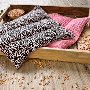Könnte beinhalten: Ein Holztablett mit drei Wärmepackungen. Zwei Packungen haben einen schwarz-beigen Leopardenmusterdruck, eine ist rosa und hat eine Waffelstruktur. Eine kleine Schüssel mit Getreide ist ebenfalls vorhanden. Getreide liegt auf der Holzoberfläche verstreut.