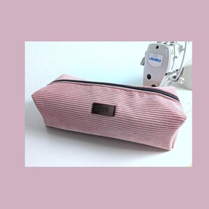 Può includere: Astuccio rosa in velluto a coste con cerniera nera e una piccola etichetta in pelle marrone. L'astuccio rettangolare è su una superficie bianca, con una macchina da cucire sullo sfondo.