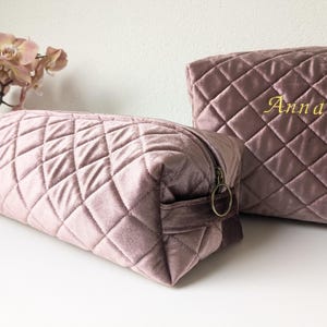 以下が含まれることがあります： 2つのモーブベルベットキルティングコスメティックバッグ。大きなバッグには「Anna」という名前が金色の刺繍で施されています。バッグはダイヤモンド柄で、ジッパー開閉です。背景にはピンクの蘭があります。