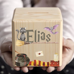 Könnte beinhalten: Ein hölzerner Sparblock mit einem Schlitz oben, verziert mit Illustrationen einer schwarzen Katze auf einem Besen, Fledermäusen und anderen magischen Elementen. Der Name "Elias" ist auf der Vorderseite aufgedruckt. Der Block wird in den Händen gehalten.