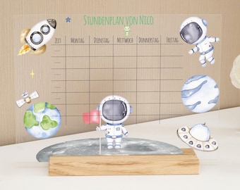 May include: A clear acrylic weekly schedule board with a space theme. The board has a grid with days of the week labeled in German: Montag, Dienstag, Mittwoch, Donnerstag, Freitag. The board is decorated with astronaut illustrations, planets, and a rocket ship.