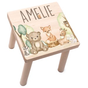 Peut inclure: Un tabouret en bois avec une finition en bois clair. Le dessus du tabouret est décoré d'un motif aquarelle d'animaux de la forêt, dont un ours, un renard, un cerf et un hibou. Le nom "Amelie" est écrit en cursive sur le dessus du tabouret.