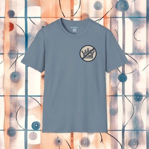 Könnte beinhalten: Hellblaues Kurzarm-T-Shirt mit einem kreisförmigen Grafik einer durchgestrichenen Krone. Die Grafik ist in Schwarz und Beige gehalten. Das Shirt hat einen Rundhalsausschnitt und ein kleines Etikett in der Nähe des Kragens.