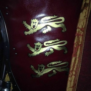 May include: A burgundy shield featuring three gold lion emblems. The shield is part of a suit of armour, with metal edging and rivets visible. A decorative banner with gold trim is on the right.