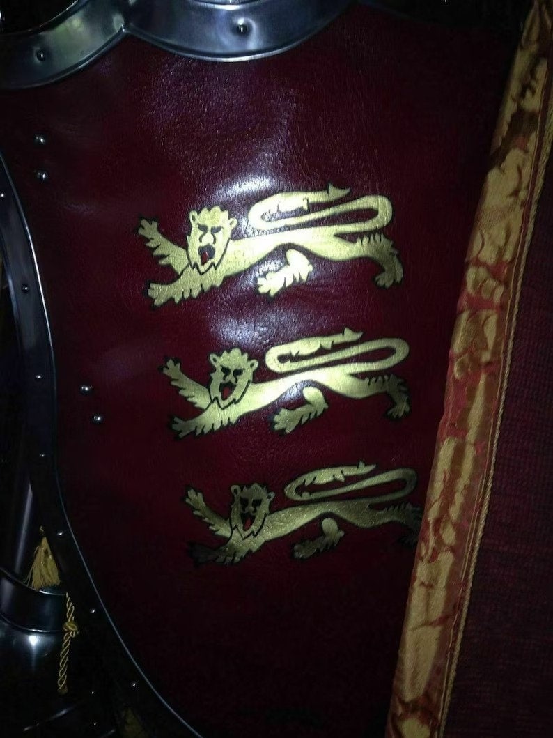 May include: A burgundy shield featuring three gold lion emblems. The shield is part of a suit of armour, with metal edging and rivets visible. A decorative banner with gold trim is on the right.