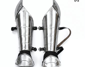 May include: Pair of polished steel greaves, part of medieval armor. The leg armor features articulated plates and leather straps for fastening. The design includes a pointed top and a curved lower section, reflecting historical craftsmanship.