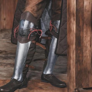 May include: A pair of metal greaves and knee cops worn over brown quilted trousers and dark brown leather shoes. The knee armour features polished steel plates secured with red leather laces, while the lower leg armour is strapped with brown leather.