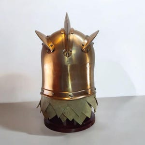 Op de afbeelding: Een messingkleurige helm met drie opvallende punten en geklonken details. De helm heeft een onderste gedeelte met overlappende, bladvormige platen. Het staat op een donkere houten standaard, met een middeleeuws of fantasiethema.