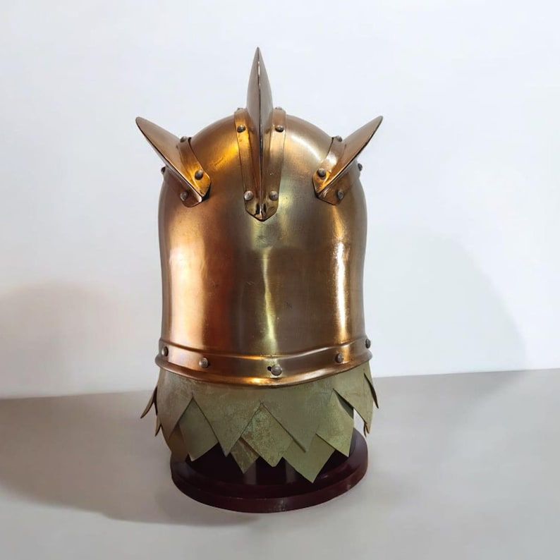 Op de afbeelding: Een messingkleurige helm met drie opvallende punten en geklonken details. De helm heeft een onderste gedeelte met overlappende, bladvormige platen. Het staat op een donkere houten standaard, met een middeleeuws of fantasiethema.