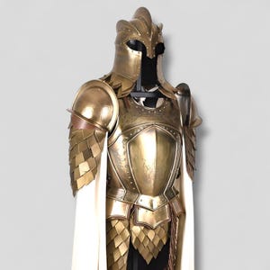 Op de afbeelding: Een gedetailleerd, goudkleurig harnas met een helm, schouderbeschermers en borstplaat. Het harnas heeft ingewikkelde ontwerpen en een witte cape. Het harnas wordt tentoongesteld op een zwarte standaard tegen een neutrale achtergrond.