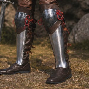 May include: Dark brown leather boots featuring polished steel greaves and knee plates. The metal armour components are secured to the footwear with brown leather straps and buckles, while the knee sections are fastened with bright red cord lacing.