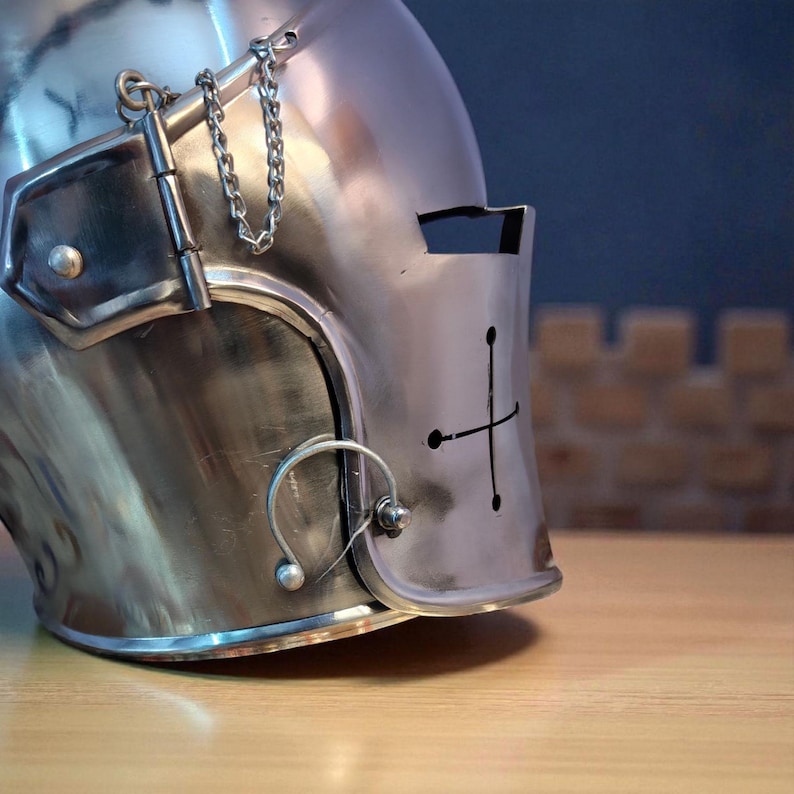 May include: Close-up of a polished, silver-colored medieval helmet. The helmet features a cross design on the front, a visor, and a chain detail. The helmet is made of metal and sits on a wooden surface.