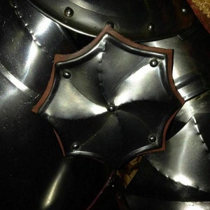 May include: Close-up of polished metal armour with a decorative, star-shaped element. The armour has a reflective surface with rivets and a brown leather trim on the star-shaped piece. The overall theme is medieval or historical.