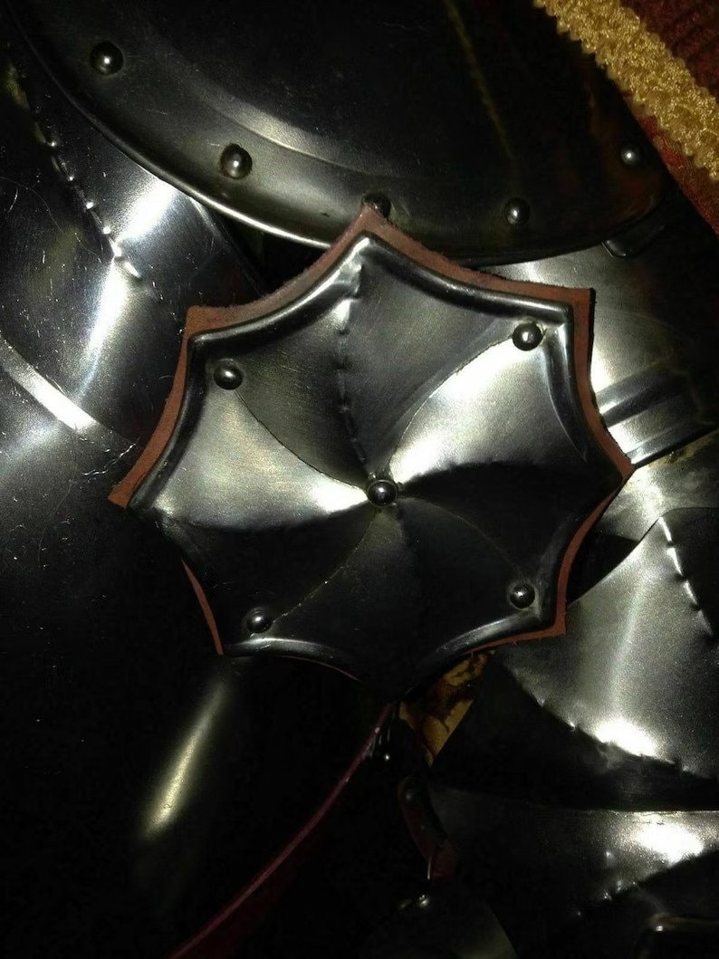 May include: Close-up of polished metal armour with a decorative, star-shaped element. The armour has a reflective surface with rivets and a brown leather trim on the star-shaped piece. The overall theme is medieval or historical.