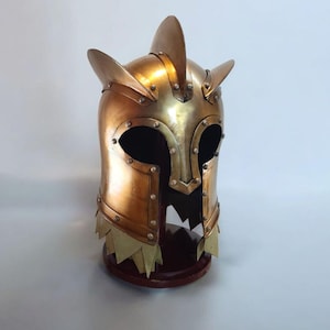 Op de afbeelding: Een goudkleurige helm in middeleeuwse stijl met puntige oorachtige uitsteeksels en een gekartelde onderrand. De helm is geklonken en lijkt van metaal te zijn. Hij staat op een donkere houten basis.