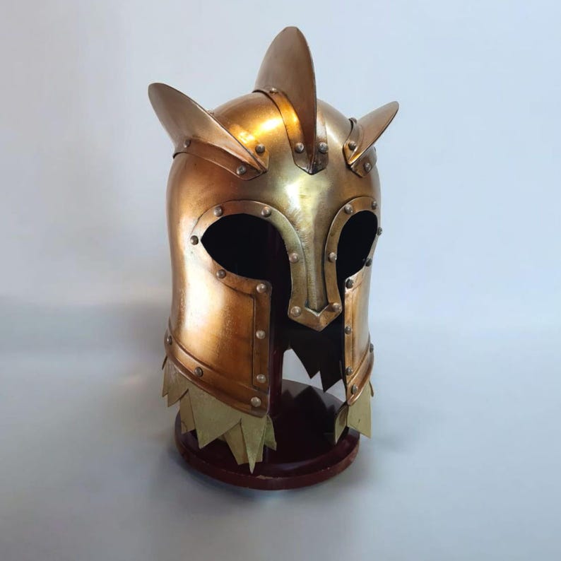 Op de afbeelding: Een goudkleurige helm in middeleeuwse stijl met puntige oorachtige uitsteeksels en een gekartelde onderrand. De helm is geklonken en lijkt van metaal te zijn. Hij staat op een donkere houten basis.