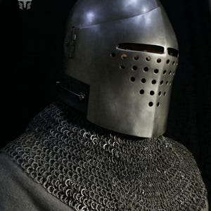 Puede incluir: Un casco de acero de estilo medieval, con una visera perforada y una cota de malla para proteger el cuello. El casco metálico se destaca sobre un fondo oscuro. La imagen resalta la artesanía de la armadura histórica.