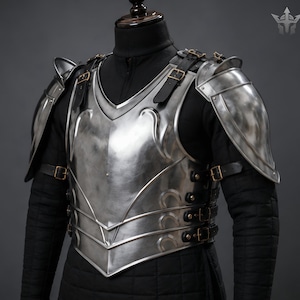 May include: A polished steel breastplate and pauldrons set featuring curved metal plates, secured with black leather straps and brass-toned buckles. The armour is worn over a black quilted long-sleeve garment, showcasing a metallic finish.