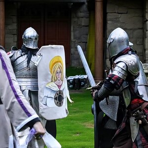 May include: Two people in medieval armor face each other, one holding a sword and the other a shield. The shield features a painting of a woman with a yellow halo. The scene takes place outdoors, with a stone building in the background.