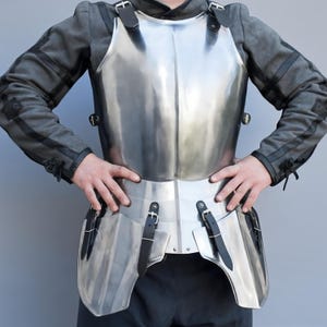 May include: A polished steel breastplate with attached waist and hip protection. The armour features black leather straps and buckles for fastening. The person is wearing a grey jacket with black accents.