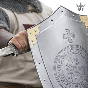 May include: A silver shield with a cross emblem and a circular seal featuring a knight on horseback. The shield has gold-colored corner accents and is held with a sword.