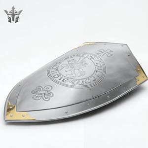 May include: A silver-colored shield with gold corner accents. The shield features an engraved emblem with a knight on horseback and a cross design. The shield has a pointed bottom and a curved top, with rivets along the edge.