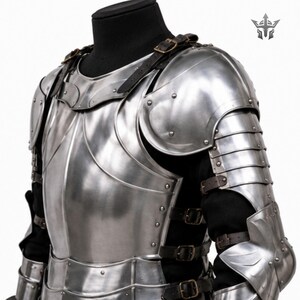 May include: A polished steel suit of armor featuring a breastplate, pauldrons, and articulated arm guards. The metal plates are secured with dark brown leather straps and brass-toned buckles over a black garment, with a logo in the top corner.