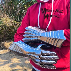 May include: A pair of silver-colored, articulated metal gauntlets, likely for medieval reenactment or costume. The gauntlets are held against a red hoodie with the text "Middle Age Medieval Zara". The metal has a polished, reflective surface.