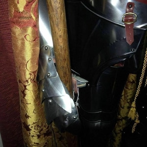 May include: Close-up of medieval armour, featuring a polished metal arm guard with riveted plates. A wooden staff is positioned next to the arm guard. The armour is black with a leather strap and gold buckle. A gold tassel hangs nearby.