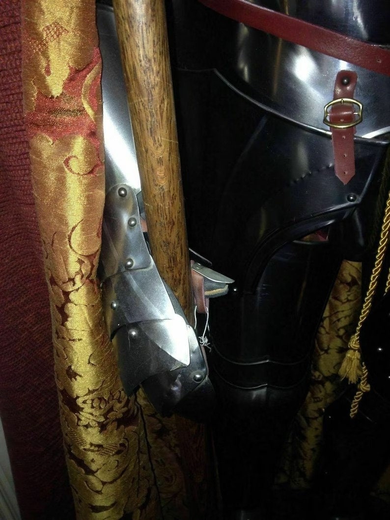 May include: Close-up of medieval armour, featuring a polished metal arm guard with riveted plates. A wooden staff is positioned next to the arm guard. The armour is black with a leather strap and gold buckle. A gold tassel hangs nearby.