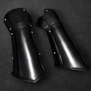 May include: A pair of black metal arm guards with a polished finish. Each guard has a curved shape, riveted details, and a flared lower edge. The arm guards are designed to protect the forearm and elbow.